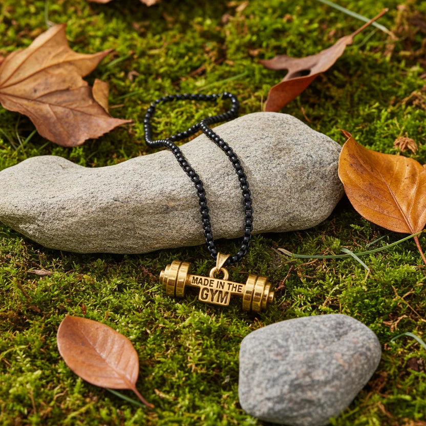 Necklace with a gold bar pendant labeled 'Made in the Gym' on a stone with moss and leaves.