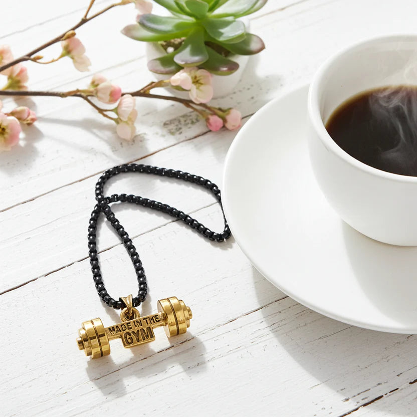Gold dumbbell necklace on a white surface with a cup of coffee and a plant.