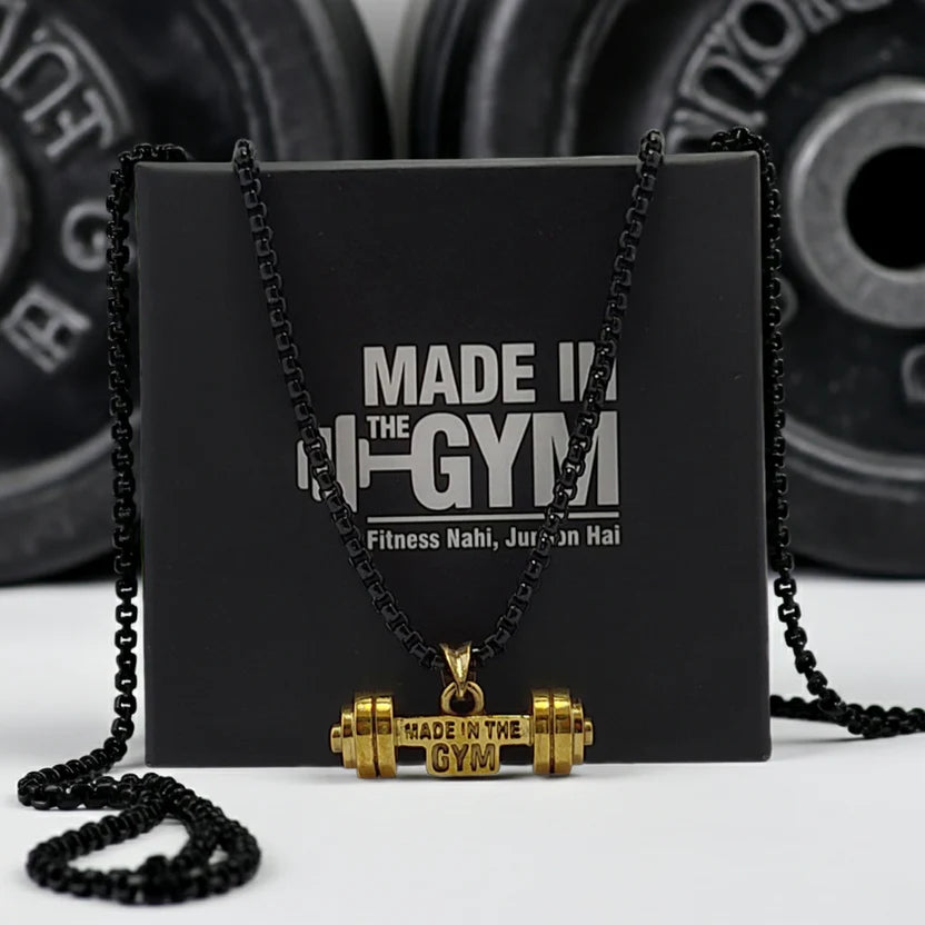 Gold dumbbell pendant necklace with 'Made in the Gym' text on a black box with gym equipment in the background.