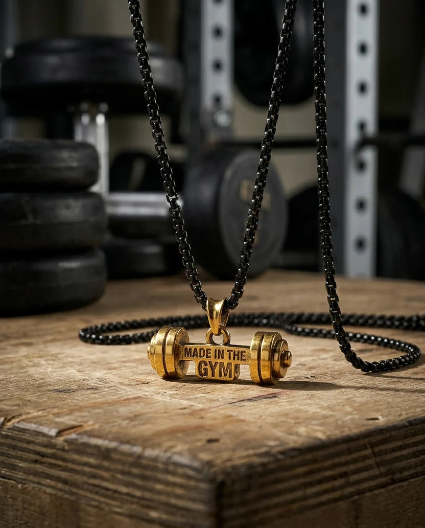 Gold barbell-shaped pendant on a black chain with 'Made in the Gym' text, placed on a wooden surface with gym equipment in the background.