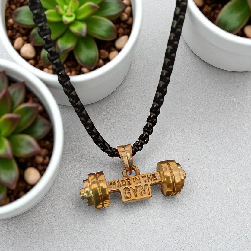Gold dumbbell pendant necklace with 'Made in the Gym' text on a black braided cord, placed on a light gray surface with potted plants.