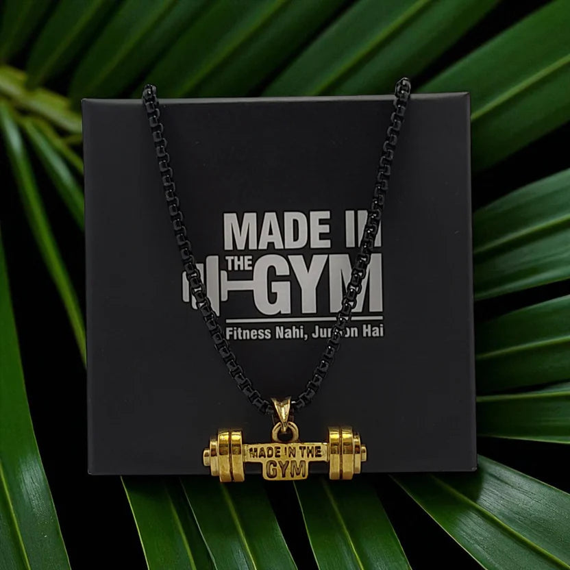 Gold barbell pendant necklace on a black card with 'Made in the Gym' text, set against a green leafy background.